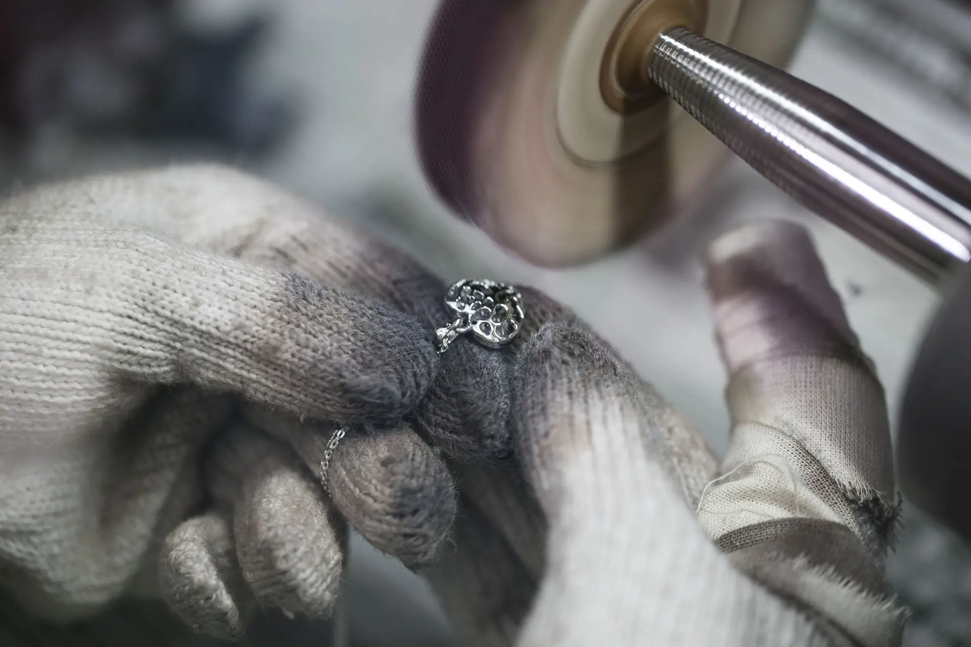 A skilled Thai artisan meticulously finishes a silver heart-shaped pendant necklace in a workshop. Close-up of gloved hands at a sanding wheel, capturing the artistry of Thailand’s silver jewellery manufacturing process.
